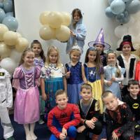 Dzieci przebrane w bajkowe stroje - górny rząd od lewej: kosmonauta, księżniczki, wróżki, pirat, król. Dolny rząd od lewej: spiderman, superbohater, batman. W tle balony żółte i niebieskie.