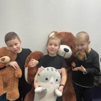 Trzech chłopców ubranych na czarno. Dwóch trzyma w rękach pluszowego misia. Chłopiec stojący na środku trzyma w ręku maskotkę kota.