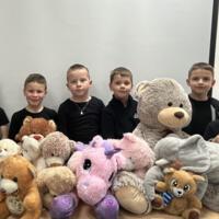 Grupa chłopców stoi. Przed nimi ułożone na stole pluszowe zabawki, miśki, słoń, jednorożec.