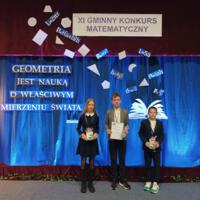 Na zdjęciu uczniowie biorący udział w konkursie ubrani na galowo. W tle na niebieskiej kotarze napis: XI Gminny Konkurs Matematyczny