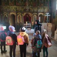 Na zdjęciu dzieci z wychowawcami wewnątrz kościoła/cerkwi. Niektóre dzieci klęczą przed krzyżem inne siedzą w ławce a jeszcze inne podziwiają ołtarz, malowidła, rzeźby.