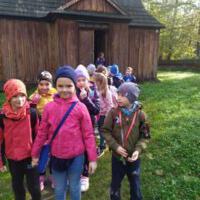 Grupa znajduje się na tle starych budynków, które są w skansenie. Dzieci są ciepło ubrane, mają na sobie kolorowe czapki i kurtki.