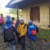 Grupa znajduje się na tle starych budynków, które są w skansenie. Dzieci są ciepło ubrane, mają na sobie kolorowe czapki i kurtki.