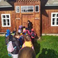 Grupa znajduje się na tle starych budynków, które są w skansenie. Dzieci są ciepło ubrane, mają na sobie kolorowe czapki i kurtki.