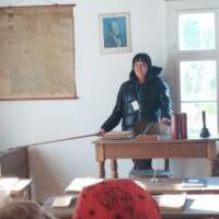 Dzieci znajdują się wewnątrz dawnej szkoły. Na ławkach uczniów są kartki do pisania i prawdziwe pióra. Pani wcieliła się w swoją rolę stając za biurkiem i uśmiechając się do dzieci. 