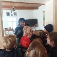 Dzieci znajdują się wewnątrz dawnej szkoły. Na ławkach uczniów są kartki do pisania i prawdziwe pióra. Pani wcieliła się w swoją rolę stając za biurkiem i uśmiechając się do dzieci. 