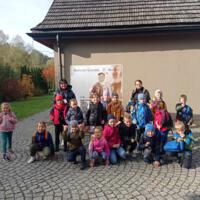 Na zdjęciu grupa dzieci z przewodnikiem i wychowawcą pozują do grupowego zdjęcia. Stoją na kostce brukowej. Za nimi biała ściana budynku oraz mały baner z napisem "Miasteczko Galicyjskie - Nowy Sącz". Z lewej strony jesienny krajobraz drzew z części ogrodu.