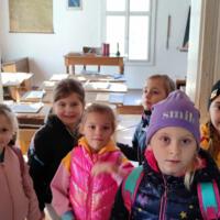 Dzieci znajdują się wewnątrz dawnej szkoły. Na ławkach uczniów są kartki do pisania i prawdziwe pióra. Pani wcieliła się w swoją rolę stając za biurkiem i uśmiechając się do dzieci. 