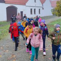 Grupa znajduje się na tle starych budynków, które są w skansenie. Dzieci są ciepło ubrane, mają na sobie kolorowe czapki i kurtki.