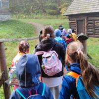 Na zdjęciach dzieci spacerują po skansenie. Trzymają się za ręce. W tle jesienna aura - drzewa z jeszcze zielonymi liśćmi na ziemi opadłe brązowe liście.