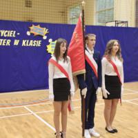 Troje uczniów - chłopiec i dwie dziewczyny ze sztandarem stoją w galowych strojach na tle dekoracji.