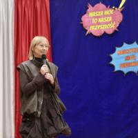 Dyrektorka szkoły ubrana w brązową sukienkę przemawia do zgromadzonych na sali uczniów, rodziców i pracowników szkoły.