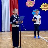 Uczniowie czytający wiersze, w tle na niebieskiej kotarze barwy narodowe oraz napis "Nauczyciel bohater w klasie"