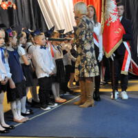 Dyrektor szkoły ubrana w sukienkę pasuje uczniów przez symboliczny gest położenia zielonego ołówka na prawym ramieniu dzieci. Na twarzach dzieci widać uśmiech. 