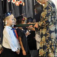 Dyrektor szkoły ubrana w sukienkę pasuje uczniów przez symboliczny gest położenia zielonego ołówka na prawym ramieniu dzieci. Na twarzach dzieci widać uśmiech. 