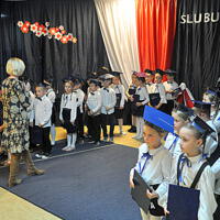 Dyrektor szkoły wygłasza mowę do mikrofonu. Mówi do uczniów oraz zebranych rodziców. Dzieci stoją i trzymają w rękach granatowe teczki.