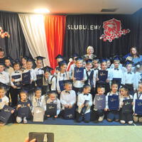 Dzieci ustawione do zdjęć grupowych z panią dyrektor oraz wychowawcami na tle dekoracji. Stoją lub kucają. Trzymają w rękach teczki z napisem "Świadectwa Szkolne". 