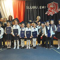 Dzieci ustawione do zdjęć grupowych z panią dyrektor oraz wychowawcami na tle dekoracji. Stoją lub kucają. Trzymają w rękach teczki z napisem "Świadectwa Szkolne". 