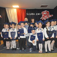 Dzieci ustawione do zdjęć grupowych z panią dyrektor oraz wychowawcami na tle dekoracji. Stoją lub kucają. Trzymają w rękach teczki z napisem "Świadectwa Szkolne". 