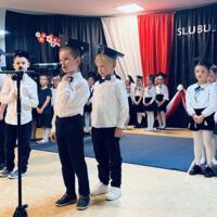 Uczniowie klas pierwszych ubrani galowo w białe koszule z granatowymi wstążkami pod szyją oraz dziewczynki granatowe lub czarne spódniczki, chłopcy granatowe lub czarne spodnie. Na głowach dzieci mają granatowe birety. W tle  na granatowej kotarze w centrum biały napis ślubujemy. Po lewej stronie barwy narodowe oraz przypięte biało czerwone kwiaty z papieru. Po prawej stronie wycięta z papieru i przymocowana do kotary głowa orła. Dzieci stoją na granatowej wykładzinie. Recytują przygotowany scenariusz.