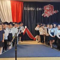 Uczniowie klas pierwszych ubrani galowo w białe koszule z granatowymi wstążkami pod szyją oraz dziewczynki granatowe lub czarne spódniczki, chłopcy granatowe lub czarne spodnie. Na głowach dzieci mają granatowe birety. W tle  na granatowej kotarze w centrum biały napis ślubujemy. Po lewej stronie barwy narodowe oraz przypięte biało czerwone kwiaty z papieru. Po prawej stronie wycięta z papieru i przymocowana do kotary głowa orła. Dzieci stoją na granatowej wykładzinie. Recytują przygotowany scenariusz.