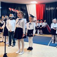 Uczniowie klas pierwszych ubrani galowo w białe koszule z granatowymi wstążkami pod szyją oraz dziewczynki granatowe lub czarne spódniczki, chłopcy granatowe lub czarne spodnie. Na głowach dzieci mają granatowe birety. W tle  na granatowej kotarze w centrum biały napis ślubujemy. Po lewej stronie barwy narodowe oraz przypięte biało czerwone kwiaty z papieru. Po prawej stronie wycięta z papieru i przymocowana do kotary głowa orła. Dzieci stoją na granatowej wykładzinie. Recytują przygotowany scenariusz.