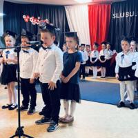 Uczniowie klas pierwszych ubrani galowo w białe koszule z granatowymi wstążkami pod szyją oraz dziewczynki granatowe lub czarne spódniczki, chłopcy granatowe lub czarne spodnie. Na głowach dzieci mają granatowe birety. W tle  na granatowej kotarze w centrum biały napis ślubujemy. Po lewej stronie barwy narodowe oraz przypięte biało czerwone kwiaty z papieru. Po prawej stronie wycięta z papieru i przymocowana do kotary głowa orła. Dzieci stoją na granatowej wykładzinie. Recytują przygotowany scenariusz.