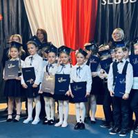 Dzieci ustawione do zdjęć grupowych z panią dyrektor oraz wychowawcami na tle dekoracji. Stoją lub kucają. Trzymają w rękach teczki z napisem "Świadectwa Szkolne". 