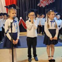 Uczniowie klas pierwszych ubrani galowo w białe koszule z granatowymi wstążkami pod szyją oraz dziewczynki granatowe lub czarne spódniczki, chłopcy granatowe lub czarne spodnie. Na głowach dzieci mają granatowe birety. W tle  na granatowej kotarze w centrum biały napis ślubujemy. Po lewej stronie barwy narodowe oraz przypięte biało czerwone kwiaty z papieru. Po prawej stronie wycięta z papieru i przymocowana do kotary głowa orła. Dzieci stoją na granatowej wykładzinie. Recytują przygotowany scenariusz.