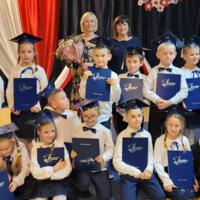 Dzieci ustawione do zdjęć grupowych z panią dyrektor oraz wychowawcami na tle dekoracji. Stoją lub kucają. Trzymają w rękach teczki z napisem "Świadectwa Szkolne". 