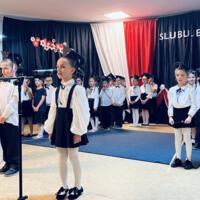 Uczniowie klas pierwszych ubrani galowo w białe koszule z granatowymi wstążkami pod szyją oraz dziewczynki granatowe lub czarne spódniczki, chłopcy granatowe lub czarne spodnie. Na głowach dzieci mają granatowe birety. W tle  na granatowej kotarze w centrum biały napis ślubujemy. Po lewej stronie barwy narodowe oraz przypięte biało czerwone kwiaty z papieru. Po prawej stronie wycięta z papieru i przymocowana do kotary głowa orła. Dzieci stoją na granatowej wykładzinie. Recytują przygotowany scenariusz.