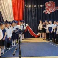 Dyrektor szkoły wygłasza mowę do mikrofonu. Mówi do uczniów oraz zebranych rodziców. Dzieci stoją i trzymają w rękach granatowe teczki.