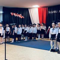 Uczniowie klas pierwszych ubrani galowo w białe koszule z granatowymi wstążkami pod szyją oraz dziewczynki granatowe lub czarne spódniczki, chłopcy granatowe lub czarne spodnie. Na głowach dzieci mają granatowe birety. W tle  na granatowej kotarze w centrum biały napis ślubujemy. Po lewej stronie barwy narodowe oraz przypięte biało czerwone kwiaty z papieru. Po prawej stronie wycięta z papieru i przymocowana do kotary głowa orła. Dzieci stoją na granatowej wykładzinie. Recytują przygotowany scenariusz.