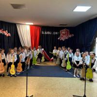 Dzieci stoją podzielenie na dwie grupy. Po prawej i lewej stronie miejsca występu, tak aby ich twarze były widoczne. W rękach trzymają wykonane z papieru żółte, zielone, pomarańczowe i brązowe gitary. W tle dekoracja. 