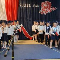 Uczniowie klas pierwszych ubrani galowo w białe koszule z granatowymi wstążkami pod szyją oraz dziewczynki granatowe lub czarne spódniczki, chłopcy granatowe lub czarne spodnie. Na głowach dzieci mają granatowe birety. W tle  na granatowej kotarze w centrum biały napis ślubujemy. Po lewej stronie barwy narodowe oraz przypięte biało czerwone kwiaty z papieru. Po prawej stronie wycięta z papieru i przymocowana do kotary głowa orła. Dzieci stoją na granatowej wykładzinie. Recytują przygotowany scenariusz.