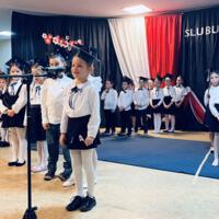 Uczniowie klas pierwszych ubrani galowo w białe koszule z granatowymi wstążkami pod szyją oraz dziewczynki granatowe lub czarne spódniczki, chłopcy granatowe lub czarne spodnie. Na głowach dzieci mają granatowe birety. W tle  na granatowej kotarze w centrum biały napis ślubujemy. Po lewej stronie barwy narodowe oraz przypięte biało czerwone kwiaty z papieru. Po prawej stronie wycięta z papieru i przymocowana do kotary głowa orła. Dzieci stoją na granatowej wykładzinie. Recytują przygotowany scenariusz.