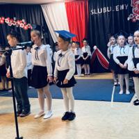 Uczniowie klas pierwszych ubrani galowo w białe koszule z granatowymi wstążkami pod szyją oraz dziewczynki granatowe lub czarne spódniczki, chłopcy granatowe lub czarne spodnie. Na głowach dzieci mają granatowe birety. W tle  na granatowej kotarze w centrum biały napis ślubujemy. Po lewej stronie barwy narodowe oraz przypięte biało czerwone kwiaty z papieru. Po prawej stronie wycięta z papieru i przymocowana do kotary głowa orła. Dzieci stoją na granatowej wykładzinie. Recytują przygotowany scenariusz.