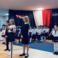 Dwie dziewczynki i chłopiec z klasy czwartej mówią do mikrofonu. Ubrani są galowo. W tle dekoracja oraz po lewej i prawej stronie pierwszoklasiści stojący za wyznaczoną na podłodze linią.