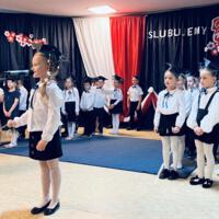 Uczniowie klas pierwszych ubrani galowo w białe koszule z granatowymi wstążkami pod szyją oraz dziewczynki granatowe lub czarne spódniczki, chłopcy granatowe lub czarne spodnie. Na głowach dzieci mają granatowe birety. W tle  na granatowej kotarze w centrum biały napis ślubujemy. Po lewej stronie barwy narodowe oraz przypięte biało czerwone kwiaty z papieru. Po prawej stronie wycięta z papieru i przymocowana do kotary głowa orła. Dzieci stoją na granatowej wykładzinie. Recytują przygotowany scenariusz.