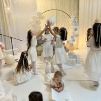 Grupa dzieci przedstawiająca jasełka. Dziewczynki ubrane na w białe sukienki i białe rajstopy, chłopcy również ubrani na biało. W tle dekoracja ze światełkami na białym materiale