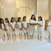 Grupa dzieci przedstawiająca jasełka. Dziewczynki ubrane na w białe sukienki i białe rajstopy, chłopcy również ubrani na biało. W tle dekoracja ze światełkami na białym materiale