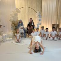 Grupa dzieci przedstawiająca jasełka. Dziewczynki ubrane na w białe sukienki i białe rajstopy, chłopcy również ubrani na biało. W tle dekoracja ze światełkami na białym materiale