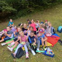 Na zdjęciach dzieci podczas spaceru po lesie, na polanie bawią się wspólnie przy użyciu chusty animacyjnej oraz bibuły w różnych kolorach. Jest słoneczna pogoda, na tle nieba pojawiają się białe chmury.