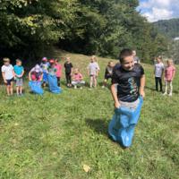 Na zdjęciach dzieci podczas spaceru po lesie, na polanie bawią się wspólnie przy użyciu chusty animacyjnej oraz bibuły w różnych kolorach. Jest słoneczna pogoda, na tle nieba pojawiają się białe chmury.
