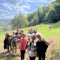 Na zdjęciach dzieci podczas spaceru po lesie, na polanie bawią się wspólnie przy użyciu chusty animacyjnej oraz bibuły w różnych kolorach. Jest słoneczna pogoda, na tle nieba pojawiają się białe chmury.