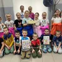Dzieci ustawione są do wspólnego zdjęcia. W centralnej części dwie dziewczynki trzymają w ręku tort urodzinowy, kilkoro dzieci ma w rękach własnoręcznie wykonane kartki urodzinowe. 