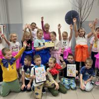 Dzieci ustawione są do wspólnego zdjęcia. W centralnej części dwie dziewczynki trzymają w ręku tort urodzinowy, kilkoro dzieci ma w rękach własnoręcznie wykonane kartki urodzinowe. 