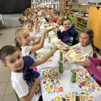 Dzieci przy wspólnym stole nakrytym białym obrusem jedzą tort urodzinowy.