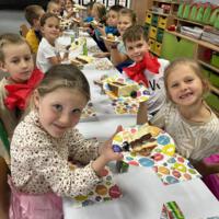 Dzieci przy wspólnym stole nakrytym białym obrusem jedzą tort urodzinowy.