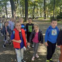 Dzieci spacerują po parku, jest słonecznie, na ziemi widać opadłe liście.